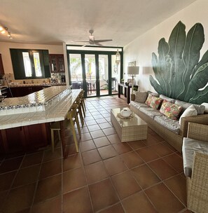 A spacious kitchen with a large island, a dining area with chairs, a sofa with cushions, and a large green leaf mural on the wall. - Spacious 3 bedroom Rio Mar Villa with Spectacular Ocean View & Golf Cart! (Río Grande)