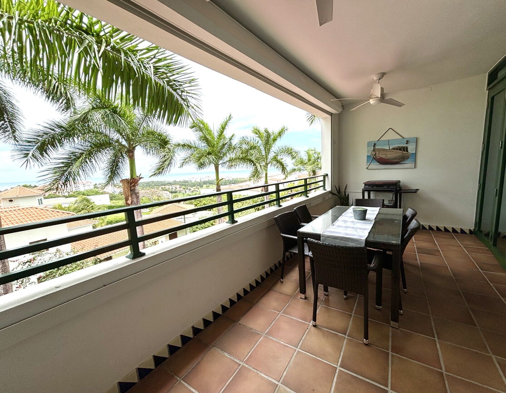 A balcony with a view of palm trees and the sea, a table with chairs, and a ceiling fan.