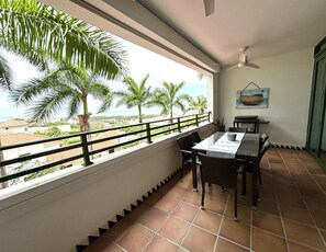 Een balkon met uitzicht op palmbomen en zee, een tafel met stoelen en een plafondventilator.