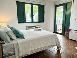 A bedroom with a bed, pillows, a bedside lamp, a window with shutters, a radiator, and a view of the outdoors. - Spacious 3 bedroom Rio Mar Villa with Spectacular Ocean View & Golf Cart! (Río Grande)