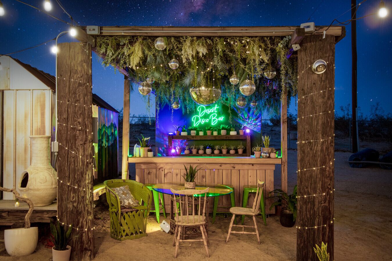A wooden outdoor bar area with a neon sign, hanging decorations, and potted plants.