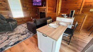 Ein gemütliches Ferienhäuschen mit einer Couch, einem kleinen Esstisch mit Tellern, einem Fernseher und einem Kamin.