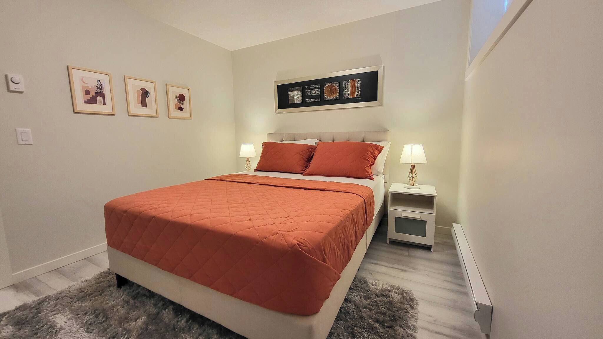 A bedroom with a bed, two bedside tables, a nightstand, a wall-mounted artwork, and a thermostat.
