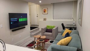 A living room with a flat-screen TV, a grey sofa, a small wooden table, and a window with blinds. - The Waterview at Rockland (Calgary)