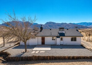 A single-story house with a gravel driveway, a wooden fence, and mountains in the background. - INTRVL Wellness House: Stargazing, Fire Pit & Desert Views (Joshua Tree)