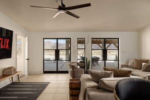 A spacious living room with a ceiling fan, a large TV, and a view of a desert landscape through the glass doors. - INTRVL Wellness House: Stargazing, Fire Pit & Desert Views (Joshua Tree)