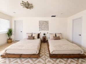A bedroom with two beds, a geometric patterned rug, a wall-mounted light fixture, and a potted plant. - INTRVL Wellness House: Stargazing, Fire Pit & Desert Views (Joshua Tree)