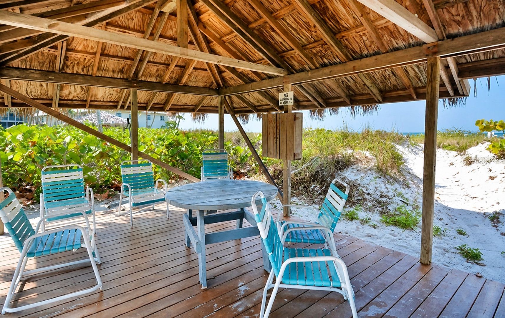A wooden deck with a thatched roof, surrounded by beach vegetation, and a table with chairs.