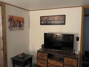 A living room with a flat-screen TV on a wooden cabinet, a small table with a duck figurine, and a wall sign that reads 'welcome to our NECK OF THE WOODS.'