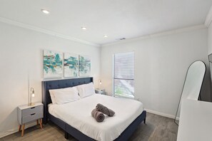 Una cama bien hecha con cabecera azul, una mesita de noche con lámpara y una obra de arte en la pared.