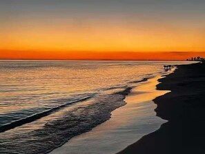 A beach at sunset with calm waves and a distant city skyline.