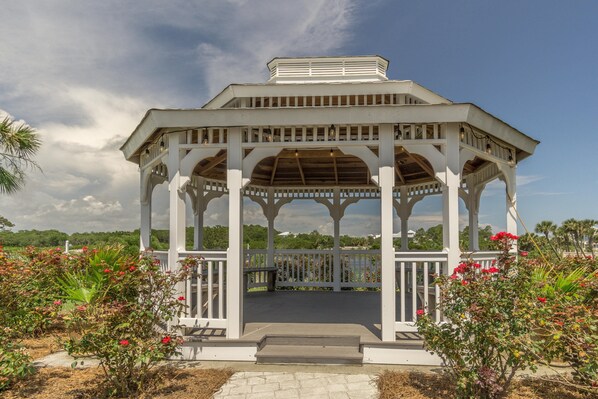 A white gazebo with a wooden roof, surrounded by flowering plants and a clear sky.