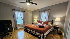 Quarto com cama, duas mesas de cabeceira com abajures, ventilador de teto e uma escrivaninha.
