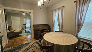 Uma sala de jantar com uma mesa redonda, cadeiras, um candeeiro e vista para uma sala de estar através de uma porta aberta.