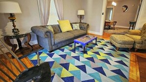 Uma sala de estar com sofá, duas poltronas, mesa de centro, tapete estampado e uma área de jantar ao fundo.