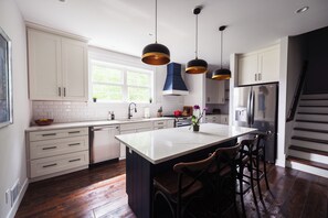 A modern kitchen with white cabinetry, a central island, pendant lighting, and a stainless steel refrigerator. -  5 Min Walk to Beach + Game Room + Wood & Propane Fire Pits - Heatonelli Haven (South Haven)