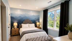 A bedroom with a bed, bedside lamps, a tufted ottoman, a wall mural, and a window with curtains. -  5 Min Walk to Beach + Game Room + Wood & Propane Fire Pits - Heatonelli Haven (South Haven)