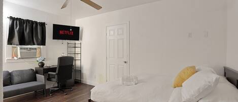 A bedroom with a bed, a desk with a chair, a ceiling fan, a TV mounted on the wall, and a door.