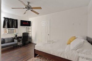 Quarto com cama, escrivaninha com cadeira, ventilador de teto, TV fixada na parede e uma porta.