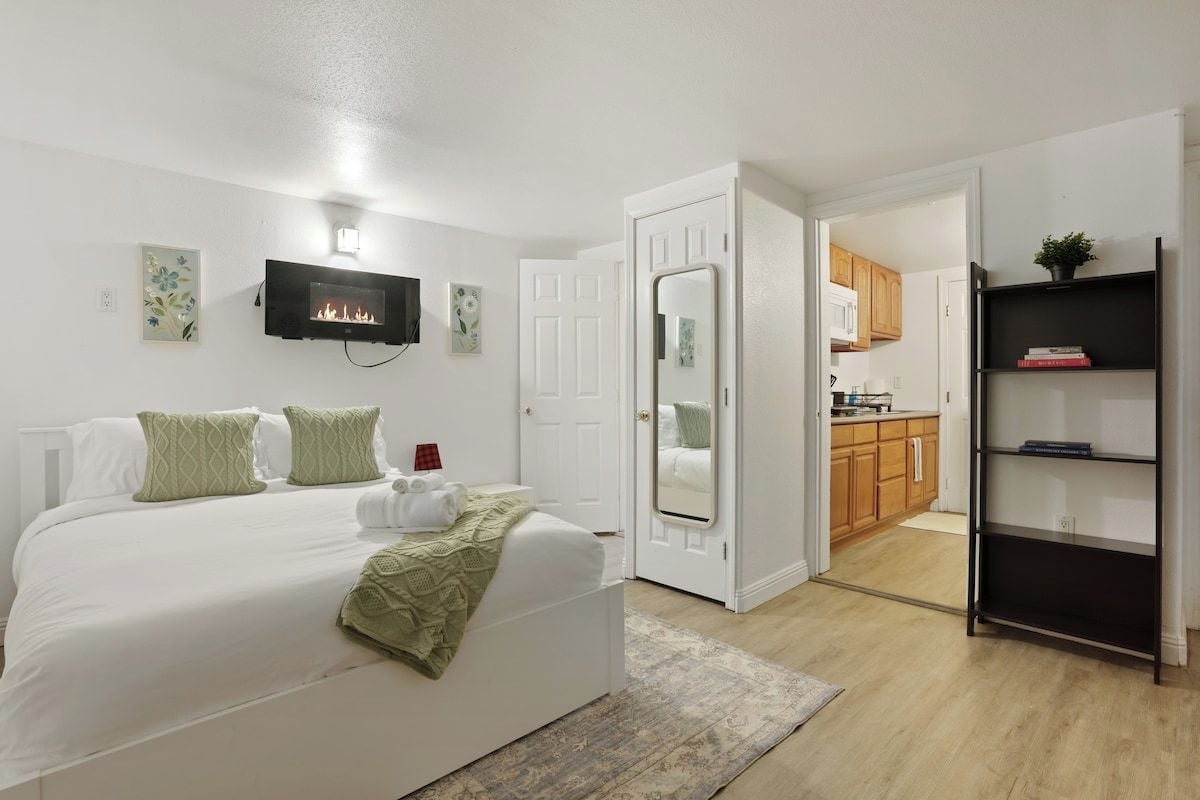 A bedroom with a bed, a fireplace, a television, and a door leading to a kitchen.