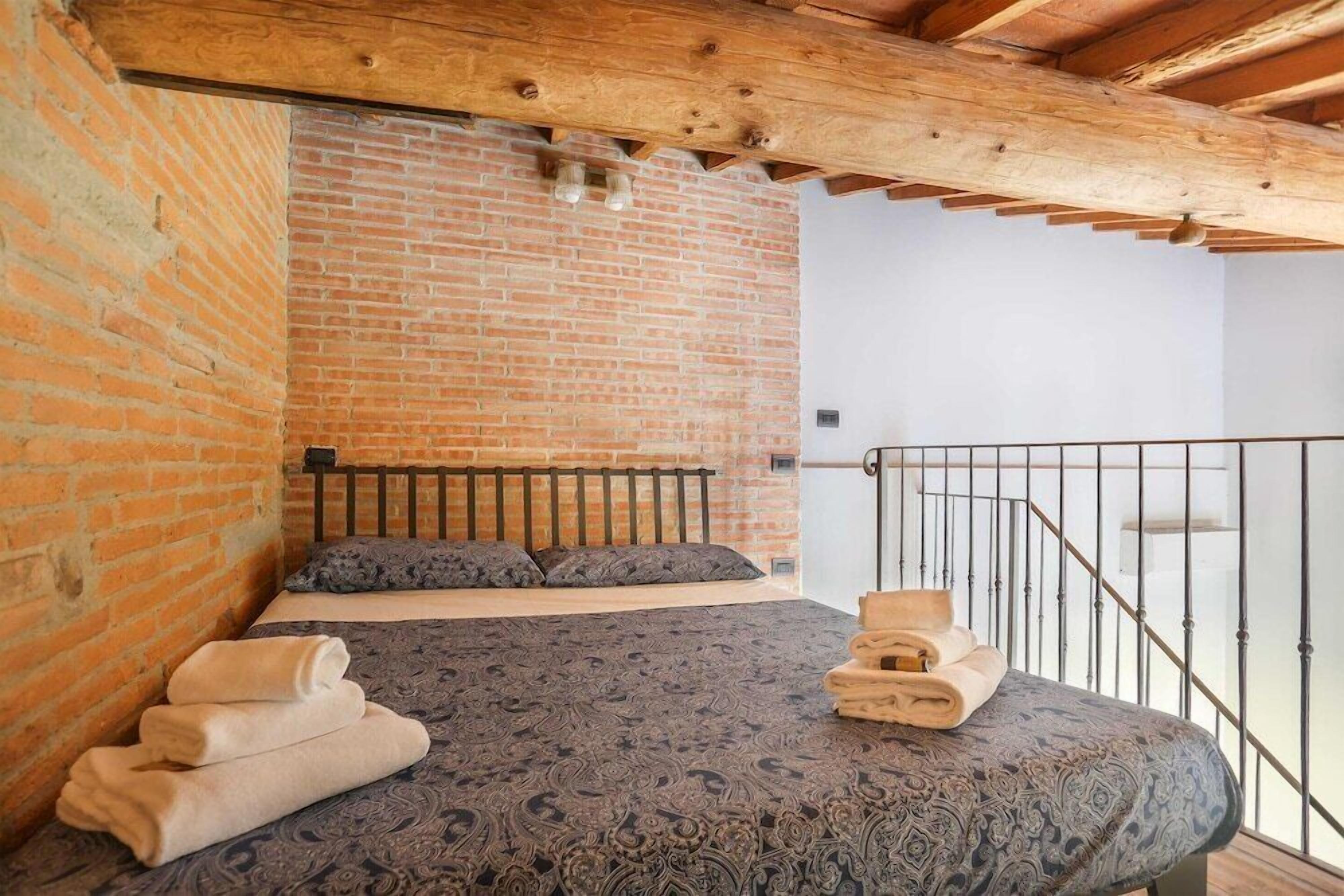 A bedroom with a brick wall, a bed with a patterned bedspread, and a balcony with a metal railing.