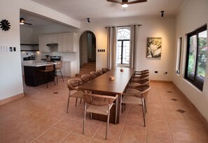 Un vaste espace à manger avec une longue table en bois et des chaises en osier, une cuisine avec des meubles blancs et une grande fenêtre donnant sur un paysage verdoyant.