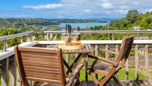 A wooden deck with a table and chairs overlooking a scenic view of a lake and hills. - Church Bay Views & Guesthouse (Waiheke Island)