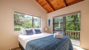 A bedroom with a bed, two windows, a wooden ceiling, and a balcony. - Church Bay Views & Guesthouse (Waiheke Island)