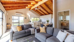 A modern living room with a grey sofa set, wooden ceiling beams, and a kitchen in the background. - Church Bay Views & Guesthouse (Waiheke Island)