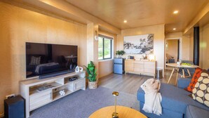 A modern living room with a flat-screen TV, a sofa, a coffee table, a small kitchen area, and a dining table. - Church Bay Views & Guesthouse (Waiheke Island)