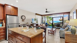 A kitchen with a large island, wooden cabinets, and a refrigerator. There is a dining area with a table and chairs, and a living room with a sofa and a view of the ocean. - Oceanfront 1BR Condo Steps to Beach West Maui (Honokowai)