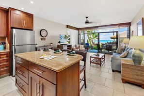 A kitchen with a large island, wooden cabinets, and a refrigerator. There is a dining area with a table and chairs, and a living room with a sofa and a view of the ocean. - Oceanfront 1BR Condo Steps to Beach West Maui (Honokowai)