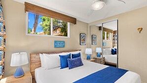 A neatly made bed with a blue accent, a wooden headboard, a nightstand with a lamp, a large mirror, and a window with a view of trees and a house. - Oceanfront 1BR Condo Steps to Beach West Maui (Honokowai)