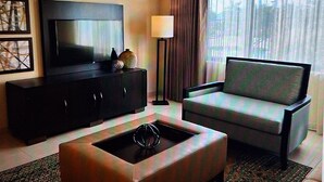 A hotel room with a flat-screen TV, a sofa, a coffee table, a lamp, and a window with curtains.