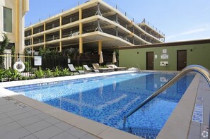 Una piscina con sillas de descanso y un edificio con aparcamiento al fondo.