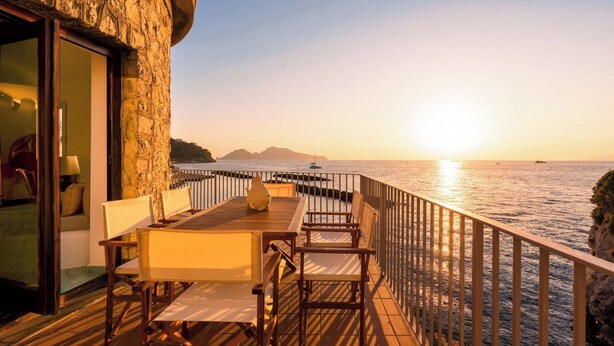 Eine Holzterrasse mit Sitzgelegenheiten im Freien, eine Steinmauer und ein Blick auf das Meer bei Sonnenuntergang.