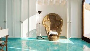 Ein Zimmer mit einem großen, verzierten Korbstuhl, einer Dreibein-Stehlampe und Blick durch ein rundbogenförmiges Fenster.