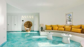 Ein Zimmer mit einem moosgrünen Fliesenboden, einem Sofa mit gelben Kissen, einem außergewöhnlichen Sessel und einem Fenster mit Blick.