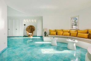 Una habitación con piso de baldosas color turquesa, un sofá con cojines amarillos, una silla singular y una ventana con vistas.
