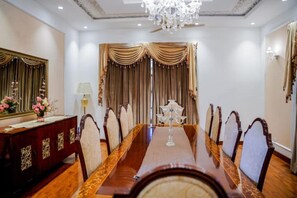 A conference room with a long table, chairs, a chandelier, curtains, a desk with a lamp, and a mirror.