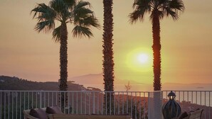Ein Balkon mit Holzstühlen und einem Tisch, mit Blick auf einen Sonnenuntergang mit Palmen und eine ferne Wasserlandschaft sowie Hügel.