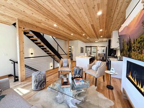 A modern living room with a glass coffee table, sofas, a fireplace, and a kitchen area in the background. - Luxe Arie Ski Haus | Amazing Mountain Views | Ski In / Ski Out | Roof Deck (Whitefish)