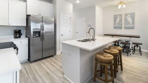 Cocina moderna con armarios blancos, electrodomésticos de acero inoxidable, una isla central con taburetes y un comedor con taburetes tipo bar.