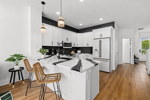 A modern kitchen with white cabinets, a central island, and a dining area with wicker chairs. - Windsor Hills Townhome - Theme Bedroom - Private Heated Pool - 2 Miles to Disney (Kissimmee)