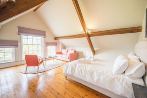 A loft bedroom with a sloped ceiling, wooden beams, a bed, a pink sofa, a chair, and large windows.