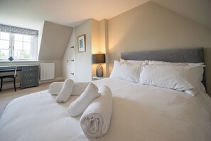 A neatly made bed with white linens and rolled towels, a desk with a chair, a lamp, and a window with blinds.