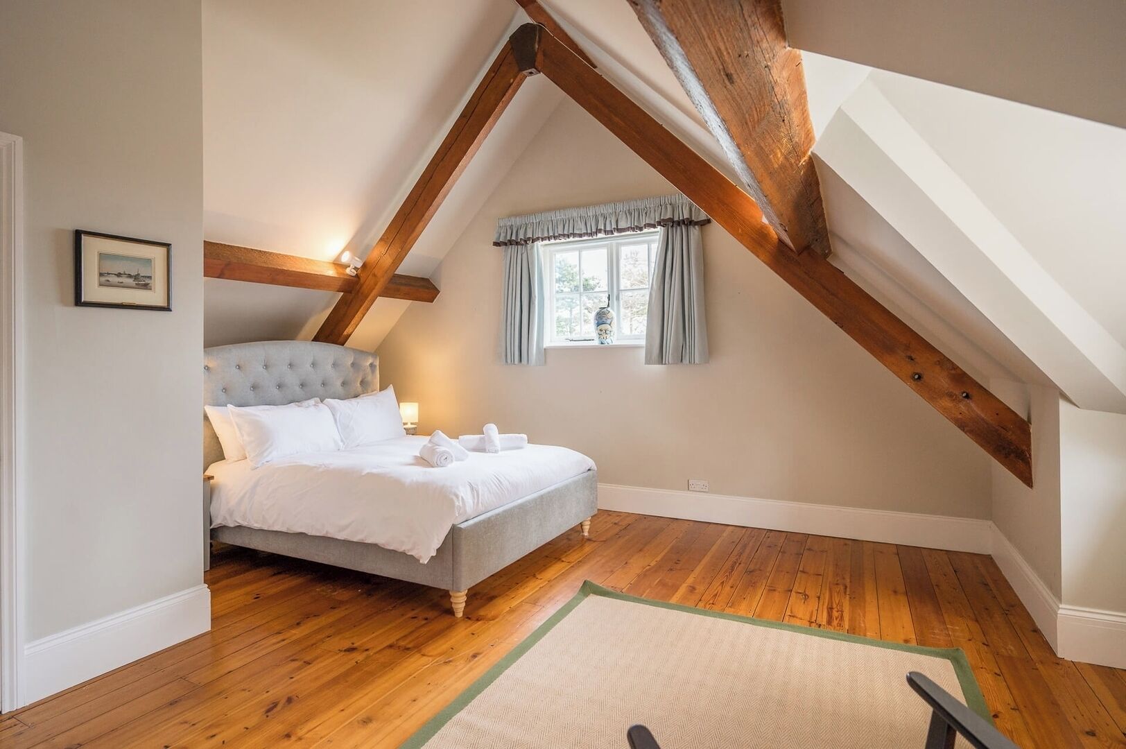 A bedroom with a large bed, wooden beams, a window with curtains, and a framed picture on the wall.
