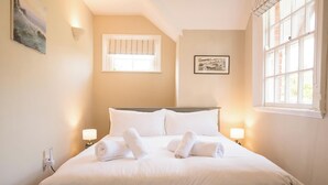 Una cama bien hecha con sábanas y toallas blancas, dos lámparas de noche, un cuadro en la pared y una ventana con persianas.