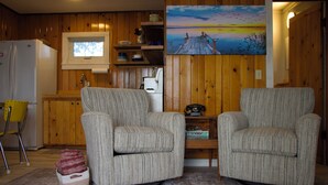 Ein Zimmer mit zwei Polstersesseln, einer Holz-Küche, einem Gemälde an der Wand und einem kleinen Tisch mit einem Telefon.