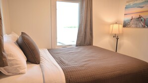 A neatly made bed with a brown quilt, a white pillow, and a white pillow on the left side. There is a window with curtains partially drawn, and a lamp on the right side of the bed.